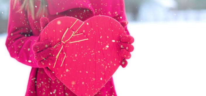 A lady in a bright pink winter outfit holding a heart-shaped bright pink box outside in the snowy weather 
